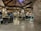 A well-lit restaurant with rustic wooden ceilings and stone pillars. There are multiple tables and chairs, some of which are empty while others are occupied by people dining. The ambiance appears cozy and inviting, with warm lighting fixtures hanging from the ceiling.