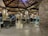 A well-lit restaurant with rustic wooden ceilings and stone pillars. There are multiple tables and chairs, some of which are empty while others are occupied by people dining. The ambiance appears cozy and inviting, with warm lighting fixtures hanging from the ceiling.