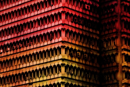 A geometric architectural façade of a high-rise building, with repetitive rectangular indentations and air conditioning units visible on the exterior. The structure is bathed in a gradient of colors ranging from red at the top to yellow at the bottom, creating a striking visual effect.