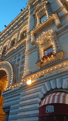 A beautifully adorned building facade embellished with numerous decorative lights highlighting its intricate architectural features. The windows are surrounded by lush floral arrangements, adding a touch of natural beauty. The structure features arches and ornate stonework, creating a sophisticated appearance, complemented by a clear evening sky.