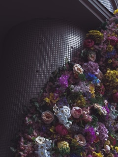 An abundance of vibrant flowers in various colors is arranged in front of a modern, textured wall. The setting appears to be indoors with a play of light and shadows accentuating the floral arrangement.