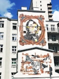 A mural on the side of a building features a large portrait within an ornate, vintage-style frame. The artwork includes multiple layers, with overlapping faces and abstract elements. Beneath, there is a collage of hands in various positions, framed with decorative patterns. The background is a light-colored building, contrasting with the vivid artwork.