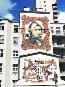 A mural on the side of a building features a large portrait within an ornate, vintage-style frame. The artwork includes multiple layers, with overlapping faces and abstract elements. Beneath, there is a collage of hands in various positions, framed with decorative patterns. The background is a light-colored building, contrasting with the vivid artwork.