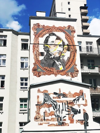 A mural on the side of a building features a large portrait within an ornate, vintage-style frame. The artwork includes multiple layers, with overlapping faces and abstract elements. Beneath, there is a collage of hands in various positions, framed with decorative patterns. The background is a light-colored building, contrasting with the vivid artwork.