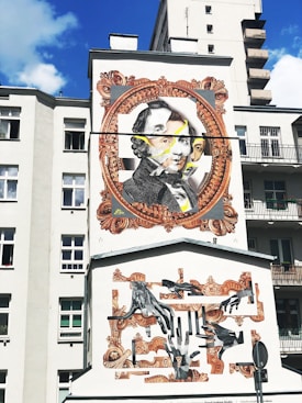 A mural on the side of a building features a large portrait within an ornate, vintage-style frame. The artwork includes multiple layers, with overlapping faces and abstract elements. Beneath, there is a collage of hands in various positions, framed with decorative patterns. The background is a light-colored building, contrasting with the vivid artwork.