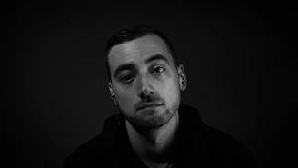 A black and white portrait of a person with short hair and a light beard wearing earphones. The subject appears to be gazing directly at the camera with a neutral expression. The background is plain and dark, which highlights the person's face and expression.