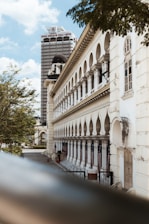 A historic building with arched windows and an ornate facade is captured in the foreground. A tall skyscraper with the name Dewan Bandaraya Kuala Lumpur is visible in the background. The scene is set under a blue sky with some scattered clouds and surrounded by green trees.