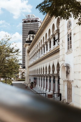 A historic building with arched windows and an ornate facade is captured in the foreground. A tall skyscraper with the name Dewan Bandaraya Kuala Lumpur is visible in the background. The scene is set under a blue sky with some scattered clouds and surrounded by green trees.