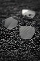 Small black triangular guitar picks with textured surfaces lie scattered among numerous tiny black plastic pellets. The focus is on the picks, with one displaying a visible logo.