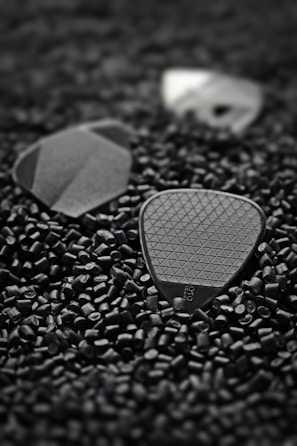 Small black triangular guitar picks with textured surfaces lie scattered among numerous tiny black plastic pellets. The focus is on the picks, with one displaying a visible logo.