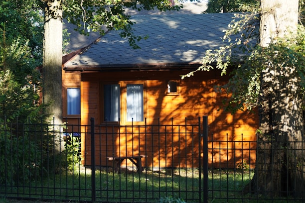 A small wooden house is surrounded by tall trees and a black metal fence. Sunlight casts shadows on the building, enhancing its warm brown color. The roof is dark gray, and the windows have blue curtains.