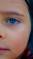 A close-up image focusing on the left eye of a person, highlighting intricate details like the blue iris and fine eyelashes. The skin appears slightly damp, possibly from water droplets.