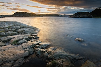 A serene coastal landscape features rugged rocks in the foreground and a calm body of water extending toward distant hills. The sky is filled with dramatic clouds and warm hues from the setting sun, casting a golden light across the scene.