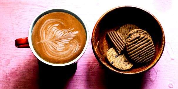A warm cup of coffee with latte art on a terracotta-colored table, surrounded by cinnamon rolls.