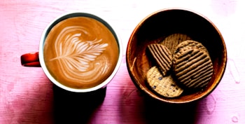 A cup of coffee with intricate latte art sits beside a wooden bowl filled with assorted cookies. The coffee is in a red mug, creating a pleasing contrast with the crema. The arrangement is placed on a pink textured background, enhancing the warm, inviting atmosphere.