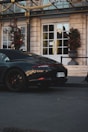 A sleek, black luxury sports car is parked on a paved street in front of a grand hotel entrance with large white doors. The area is adorned with ornate gold accents and tall potted plants with red flowers.
