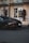 A sleek, black luxury sports car is parked on a paved street in front of a grand hotel entrance with large white doors. The area is adorned with ornate gold accents and tall potted plants with red flowers.