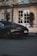 A sleek, black luxury sports car is parked on a paved street in front of a grand hotel entrance with large white doors. The area is adorned with ornate gold accents and tall potted plants with red flowers.