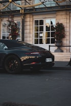A sleek, black luxury sports car is parked on a paved street in front of a grand hotel entrance with large white doors. The area is adorned with ornate gold accents and tall potted plants with red flowers.