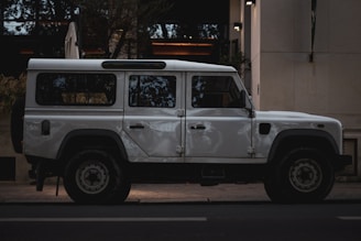 A white, rugged off-road vehicle is parked on the street next to a building. The vehicle has a boxy design with large wheels and appears sturdy, suited for adventures or expeditions.