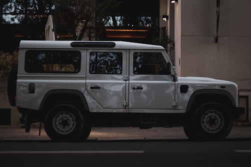 A white, rugged off-road vehicle is parked on the street next to a building. The vehicle has a boxy design with large wheels and appears sturdy, suited for adventures or expeditions.