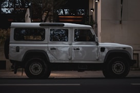 A white, rugged off-road vehicle is parked on the street next to a building. The vehicle has a boxy design with large wheels and appears sturdy, suited for adventures or expeditions.