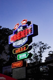 A brightly lit neon sign with multiple colors advertising a bar and restaurant. The sign features words like 'BREXIT', 'BAR-B-Q', 'BEER', and 'STEAKS' in bold, glowing letters. It's positioned against a dusky sky with silhouettes of trees in the background, creating a vibrant contrast.