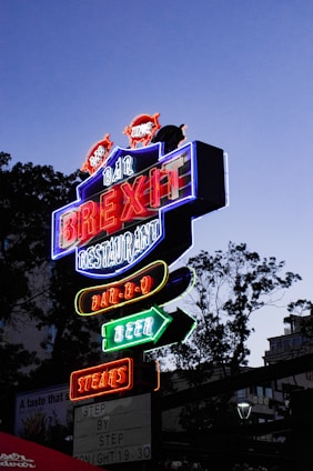 A brightly lit neon sign with multiple colors advertising a bar and restaurant. The sign features words like 'BREXIT', 'BAR-B-Q', 'BEER', and 'STEAKS' in bold, glowing letters. It's positioned against a dusky sky with silhouettes of trees in the background, creating a vibrant contrast.