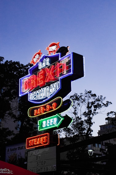 A brightly lit neon sign with multiple colors advertising a bar and restaurant. The sign features words like 'BREXIT', 'BAR-B-Q', 'BEER', and 'STEAKS' in bold, glowing letters. It's positioned against a dusky sky with silhouettes of trees in the background, creating a vibrant contrast.