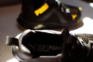 A close-up of a black athletic shoe with the word 'SOFTFOAM+' visible on the insole, suggesting cushioning and comfort. Another similar shoe is partially visible in the background, with a notable yellow logo.
