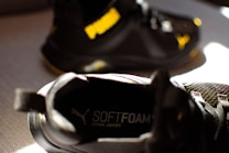 A close-up of a black athletic shoe with the word 'SOFTFOAM+' visible on the insole, suggesting cushioning and comfort. Another similar shoe is partially visible in the background, with a notable yellow logo.