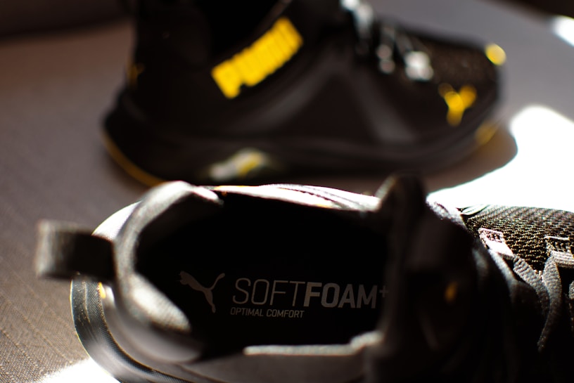 A close-up of a black athletic shoe with the word 'SOFTFOAM+' visible on the insole, suggesting cushioning and comfort. Another similar shoe is partially visible in the background, with a notable yellow logo.
