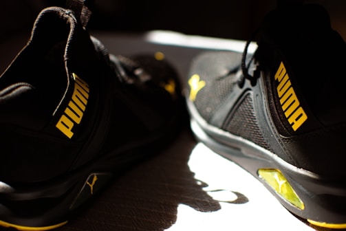 A pair of black athletic shoes with bright yellow accents and branding displayed prominently on the side. The shoes are positioned on a surface partially illuminated by sunlight, emphasizing the contrast between the colors.
