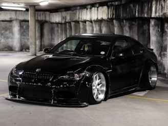 A sleek, black sports car with a low profile and custom body kit is parked in a dimly lit concrete parking garage. The car features large, shiny silver alloy wheels and tinted windows. The environment is industrial with rough, grey concrete walls and fluorescent overhead lighting.
