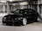 A sleek, black sports car with a low profile and custom body kit is parked in a dimly lit concrete parking garage. The car features large, shiny silver alloy wheels and tinted windows. The environment is industrial with rough, grey concrete walls and fluorescent overhead lighting.