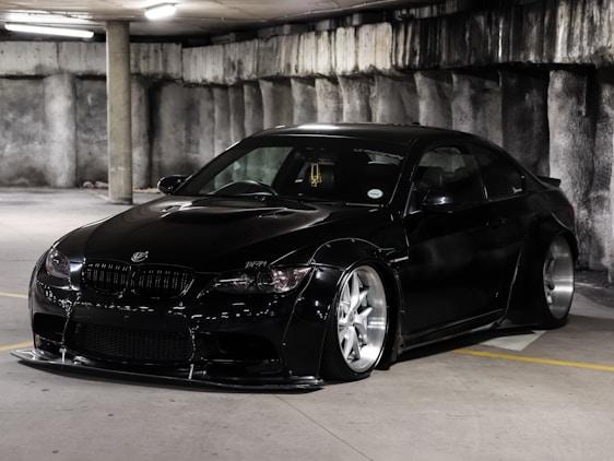 A sleek, black sports car with a low profile and custom body kit is parked in a dimly lit concrete parking garage. The car features large, shiny silver alloy wheels and tinted windows. The environment is industrial with rough, grey concrete walls and fluorescent overhead lighting.