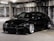 A sleek, black sports car with a low profile and custom body kit is parked in a dimly lit concrete parking garage. The car features large, shiny silver alloy wheels and tinted windows. The environment is industrial with rough, grey concrete walls and fluorescent overhead lighting.