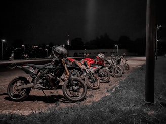 Four motorcycles are parked in a row in an outdoor area at night, illuminated by nearby streetlights. The setting is dark with minimal background details, suggesting a quiet or isolated location.