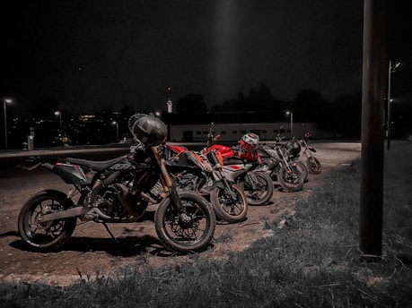 Four motorcycles are parked in a row in an outdoor area at night, illuminated by nearby streetlights. The setting is dark with minimal background details, suggesting a quiet or isolated location.