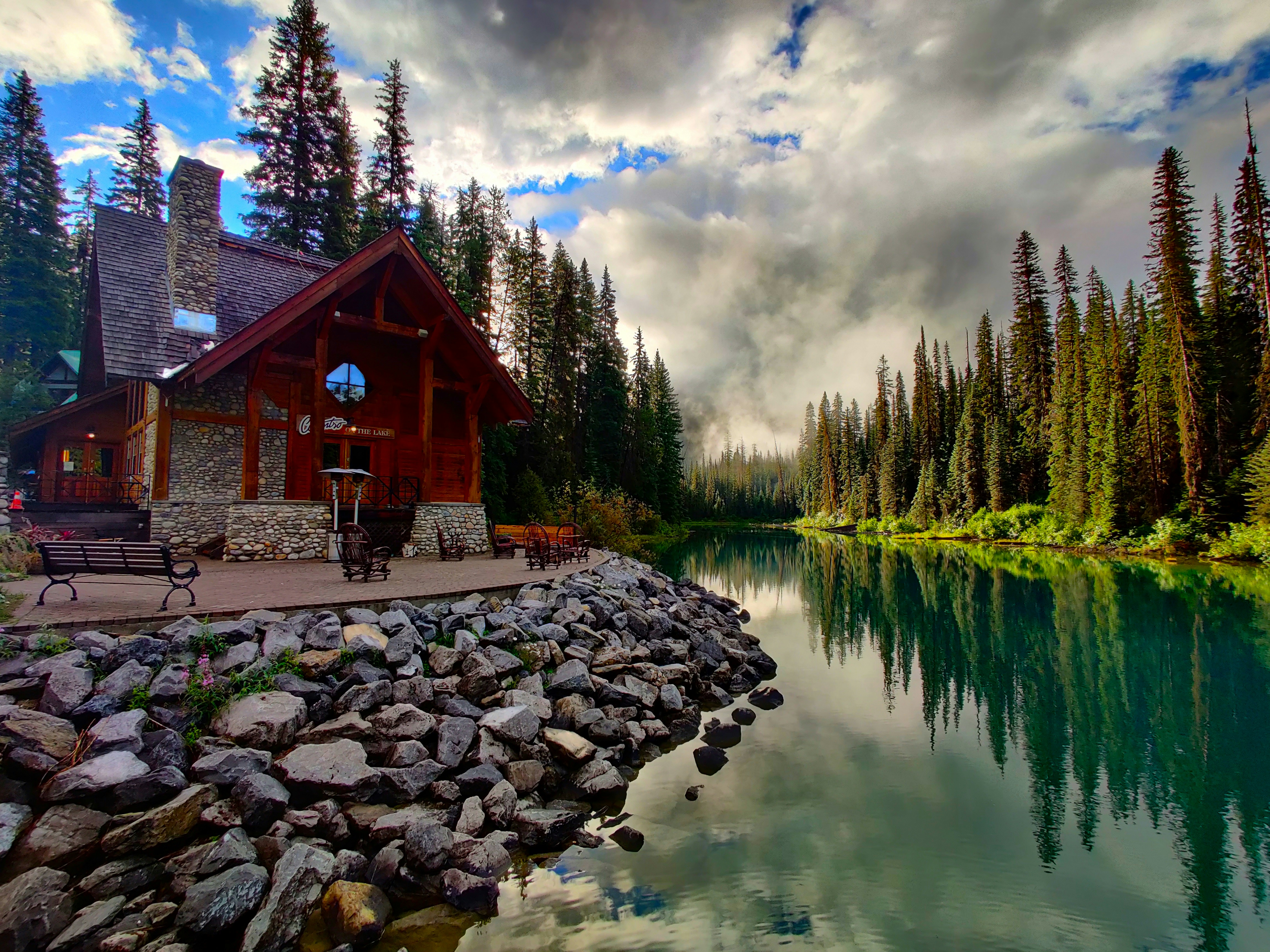 brown wooden house near green trees and river under white clouds and blue sky during daytime