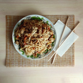 A plate of noodles with pieces of chicken and vegetables is garnished with sesame seeds. The plate is decorated with a cartoon design and is placed on a woven placemat. Beside the plate, there are chopsticks and a folded napkin.