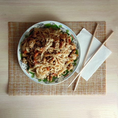 A plate of noodles with pieces of chicken and vegetables is garnished with sesame seeds. The plate is decorated with a cartoon design and is placed on a woven placemat. Beside the plate, there are chopsticks and a folded napkin.