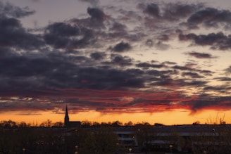 Dark clouds hover over a horizon with an orange and red sunset glow. A silhouette of a church steeple is visible in the distance against the dramatic sky.