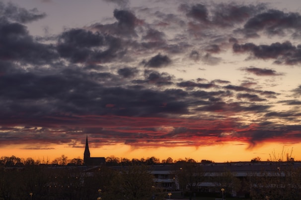 Dark clouds hover over a horizon with an orange and red sunset glow. A silhouette of a church steeple is visible in the distance against the dramatic sky.