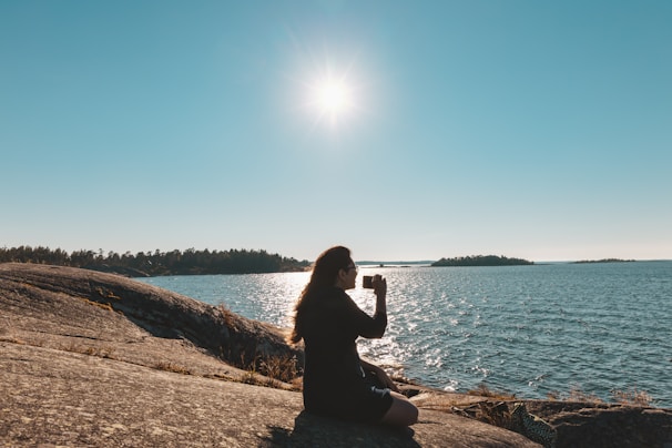 A person sits on a rocky shore, holding a camera up to their face, capturing the scenic view. The sun is shining brightly in a clear blue sky, reflecting off the water. In the distance, small islands are visible across the shimmering sea.