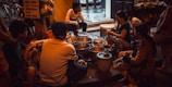 A group of people are gathered around a variety of pots and containers, suggesting a street food setting. The scene takes place in an urban environment with visible bicycles and a partially readable sign in the background. The lighting is dim and warm, creating an intimate atmosphere.