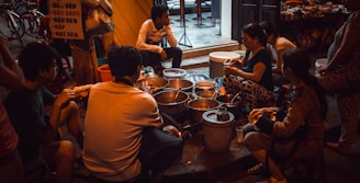 A group of people are gathered around a variety of pots and containers, suggesting a street food setting. The scene takes place in an urban environment with visible bicycles and a partially readable sign in the background. The lighting is dim and warm, creating an intimate atmosphere.