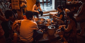A group of people are gathered around a variety of pots and containers, suggesting a street food setting. The scene takes place in an urban environment with visible bicycles and a partially readable sign in the background. The lighting is dim and warm, creating an intimate atmosphere.