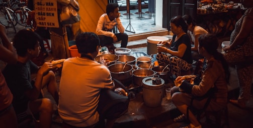 A group of people are gathered around a variety of pots and containers, suggesting a street food setting. The scene takes place in an urban environment with visible bicycles and a partially readable sign in the background. The lighting is dim and warm, creating an intimate atmosphere.