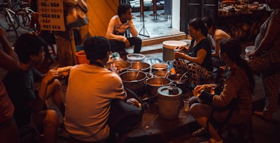 A group of people are gathered around a variety of pots and containers, suggesting a street food setting. The scene takes place in an urban environment with visible bicycles and a partially readable sign in the background. The lighting is dim and warm, creating an intimate atmosphere.